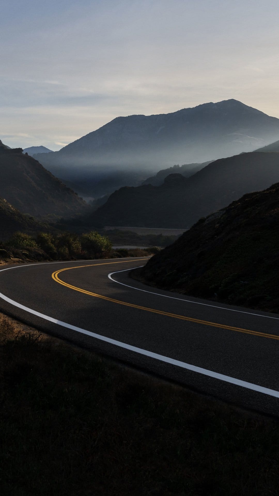 Curving asphalt road with yellow lines winds through hazy blue mountain peaks under soft morning golden sunlight.