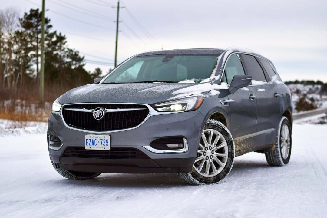Gray Buick Enclave SUV parked on a snow-covered rural road with a winter forest background under an overcast sky.