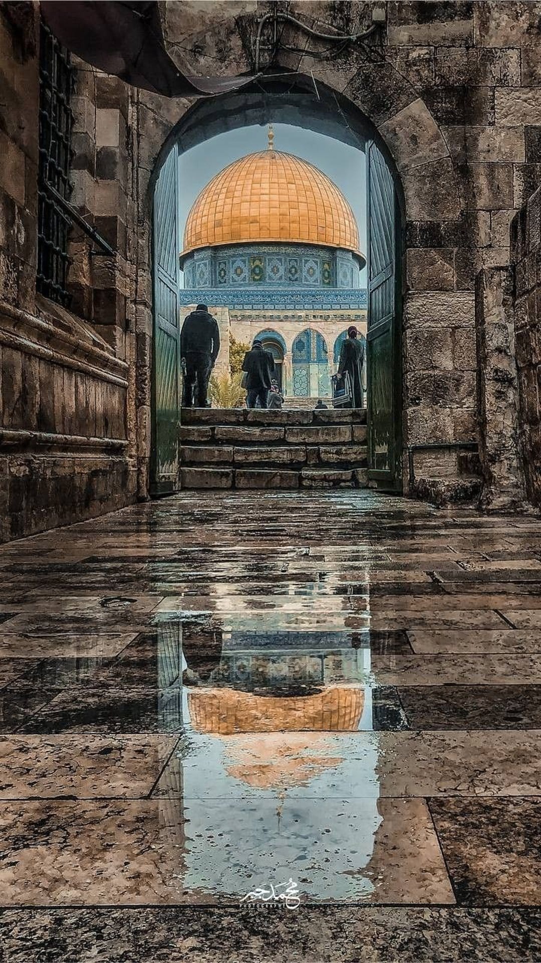 Golden Dome of the Rock reflects in a ground puddle framed by a weathered stone archway with distant people.