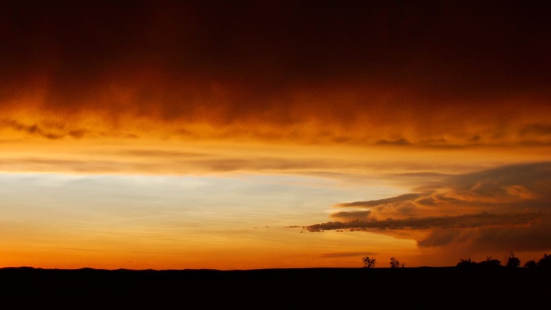 Fiery orange and red clouds swirl above a dark silhouetted horizon line under a pale blue twilight sky.