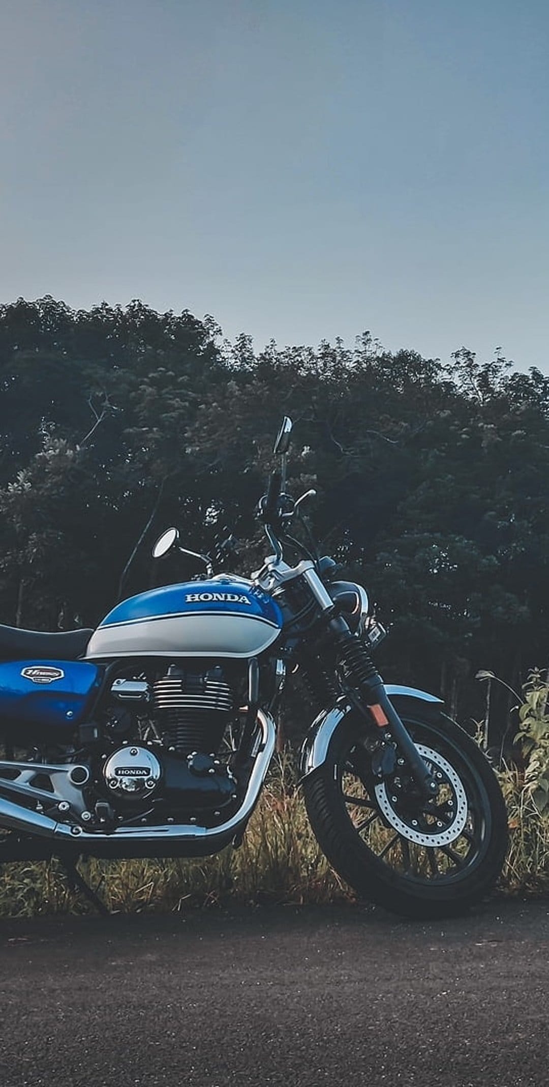 Blue Honda cruiser motorcycle parked on a paved road with green trees and a bright sky in the background.