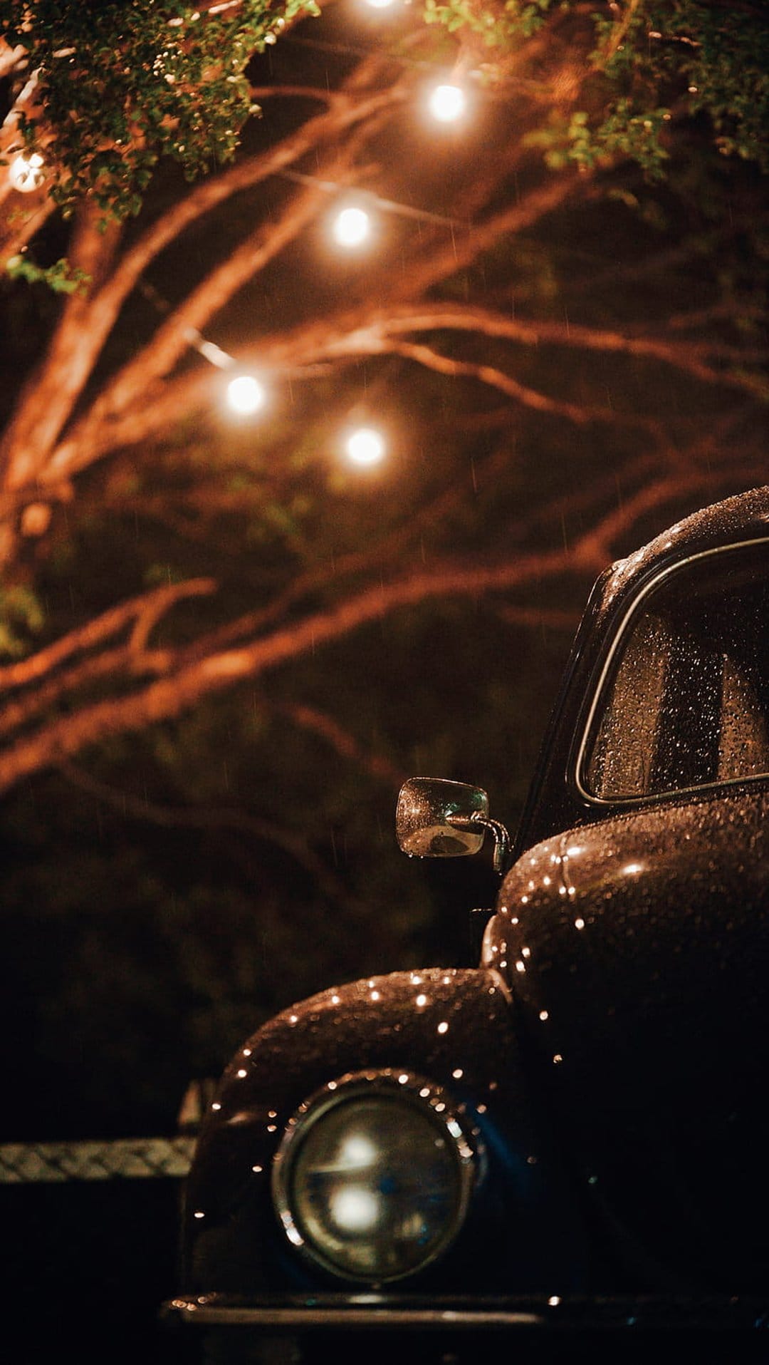 Vintage car headlight and chrome fender bead with rain under warm glowing string lights during a moody rainy night.