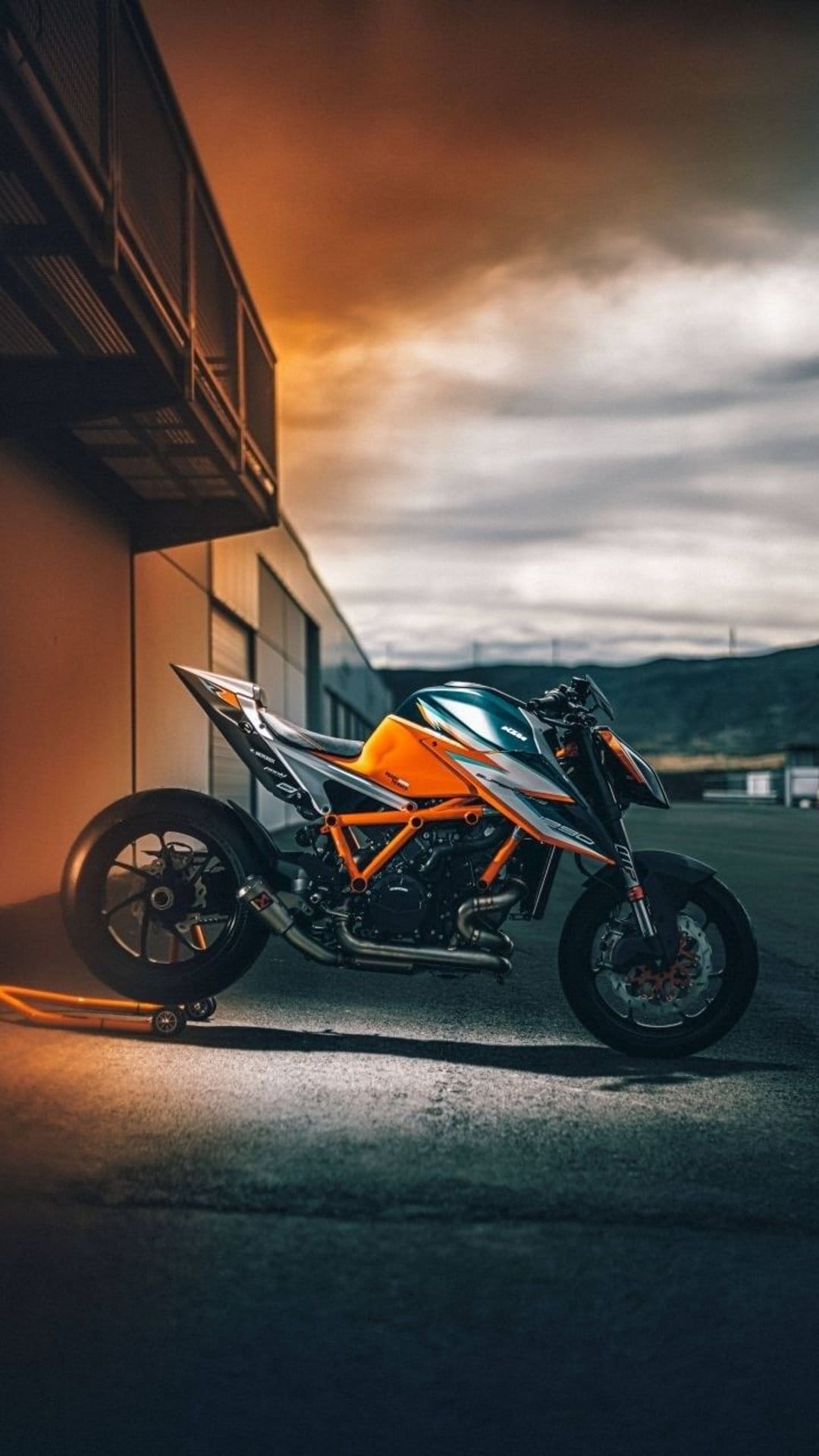 KTM motorcycle with an orange frame parked on asphalt against a backdrop of mountains and a sunset cloudy sky.
