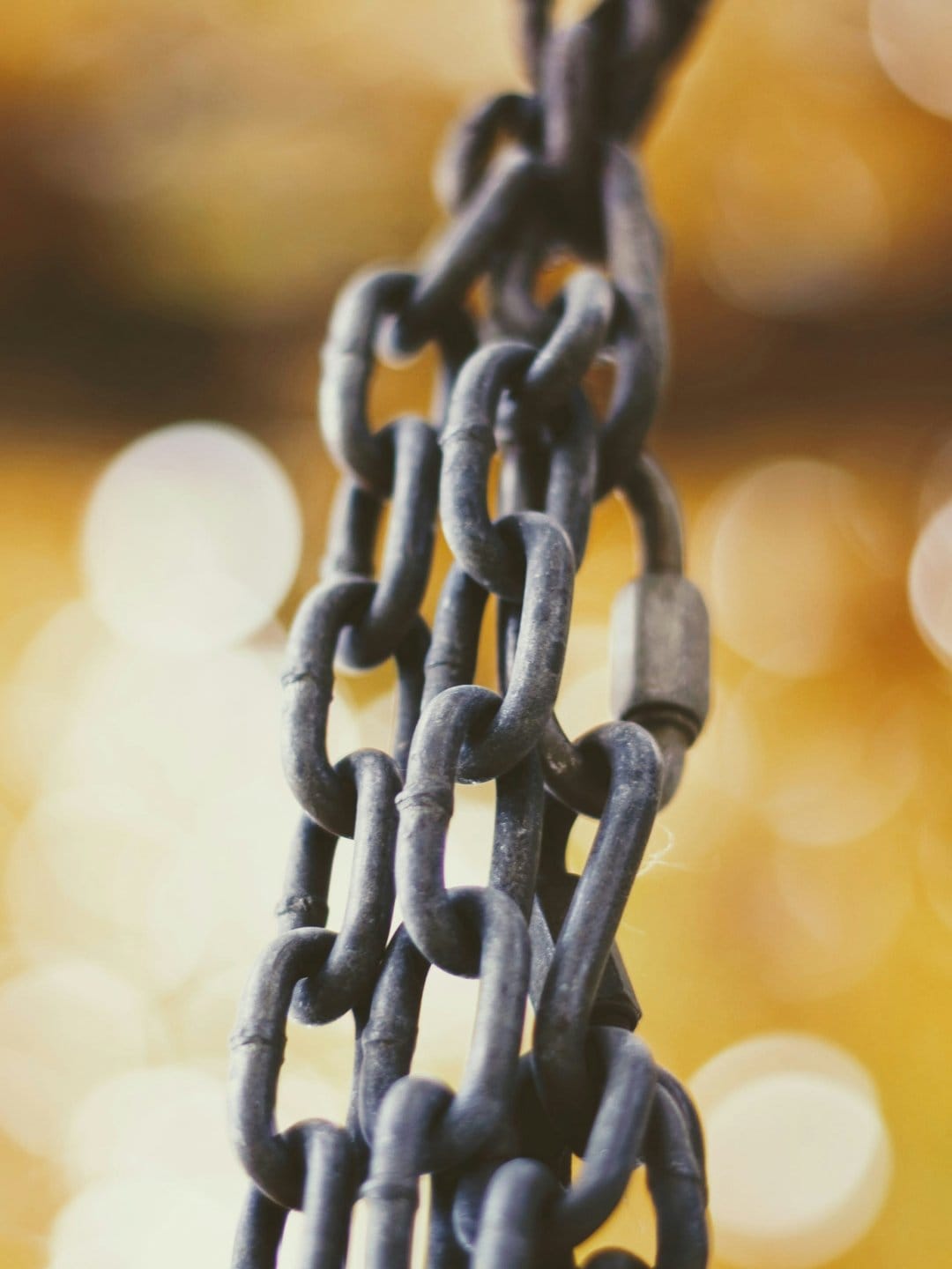 Dark weathered metal chain links hanging vertically with sharp focus against a soft golden bokeh background.