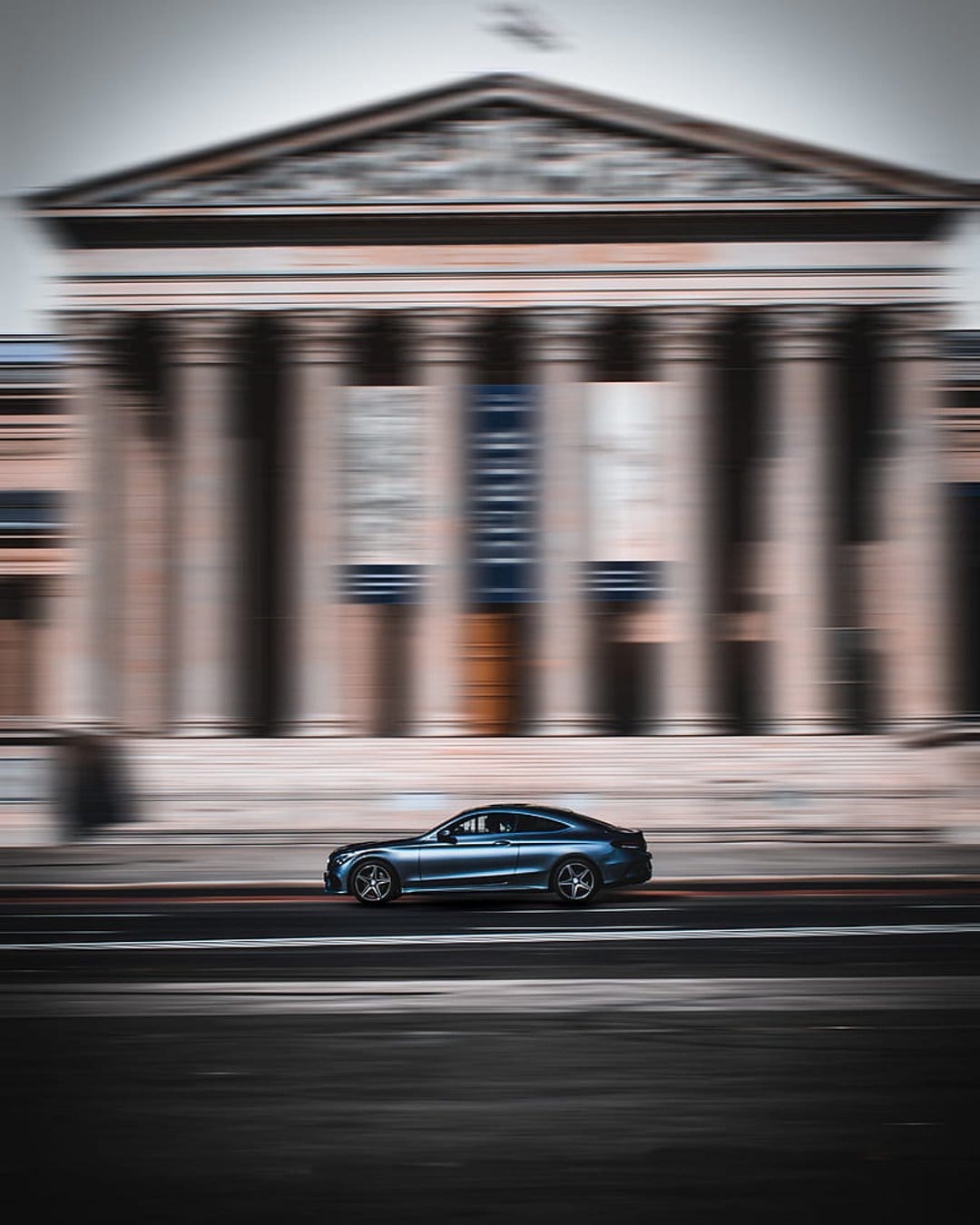 Blue coupe car speeds past a classical building with tall columns using a heavy motion blur effect on the road.