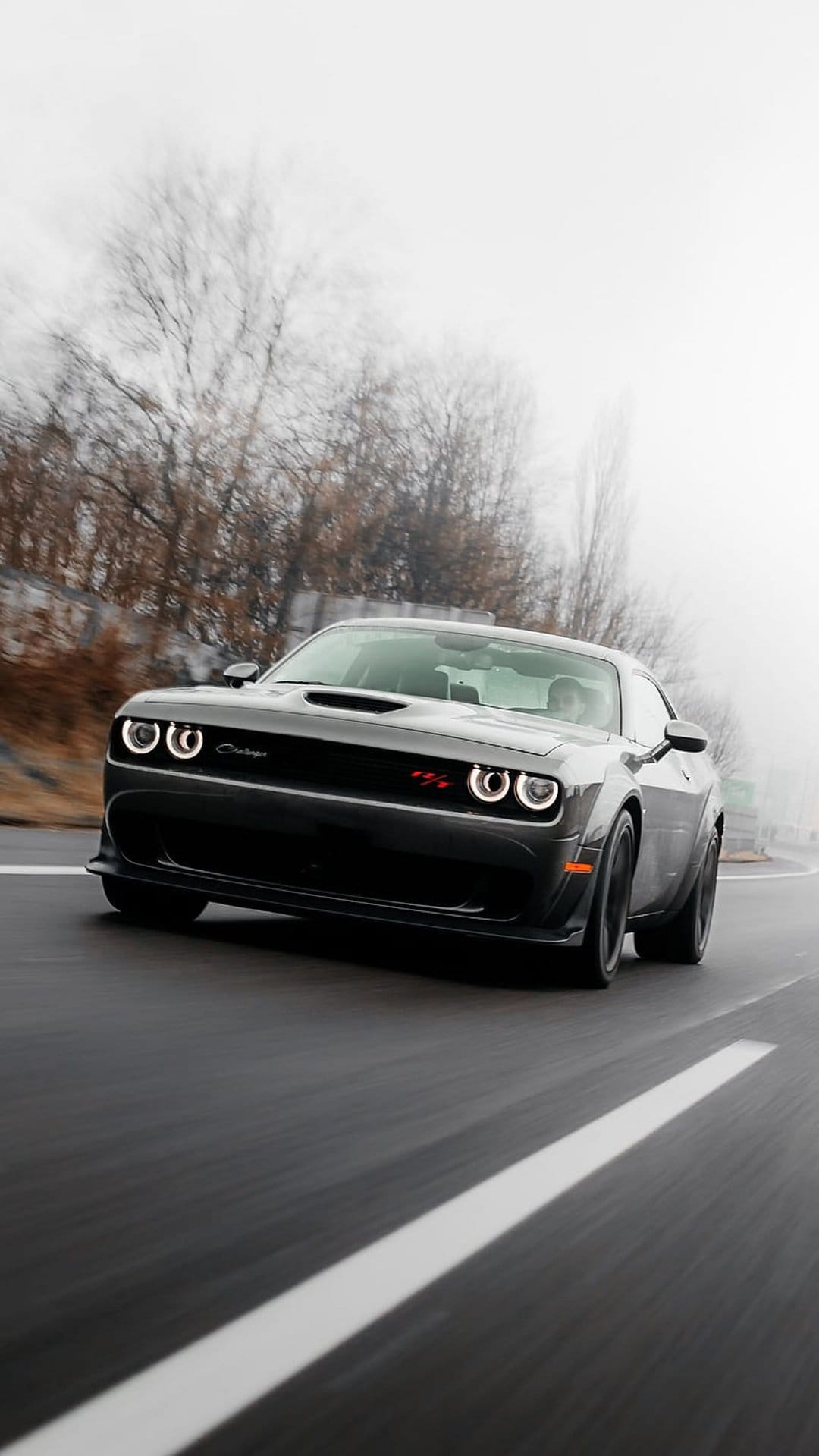 Dodge Challenger SRT Hellcat with glowing headlights driving on a wet, foggy road with blurred trees.