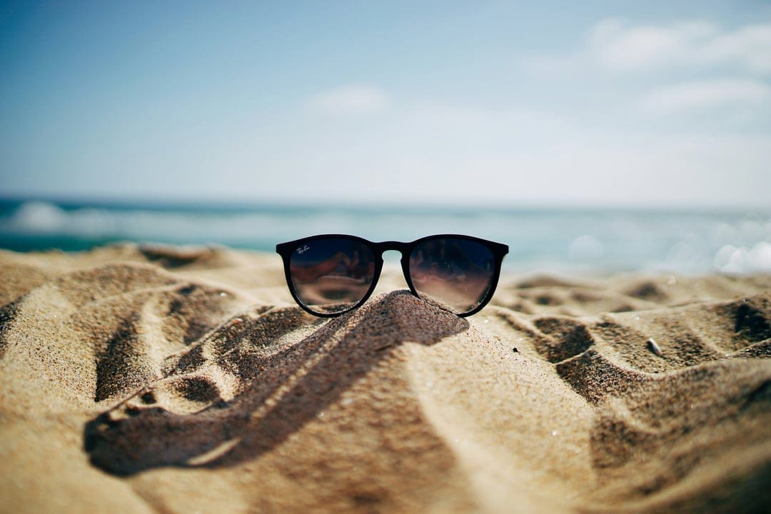 Dark sunglasses resting on rippled beach sand with soft waves and a blue sky blurred in the background.