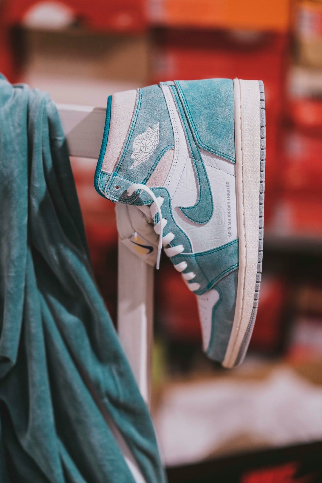 Air Jordan 1 High OG sneaker featuring aqua teal suede and white leather in front of blurred shoeboxes.
