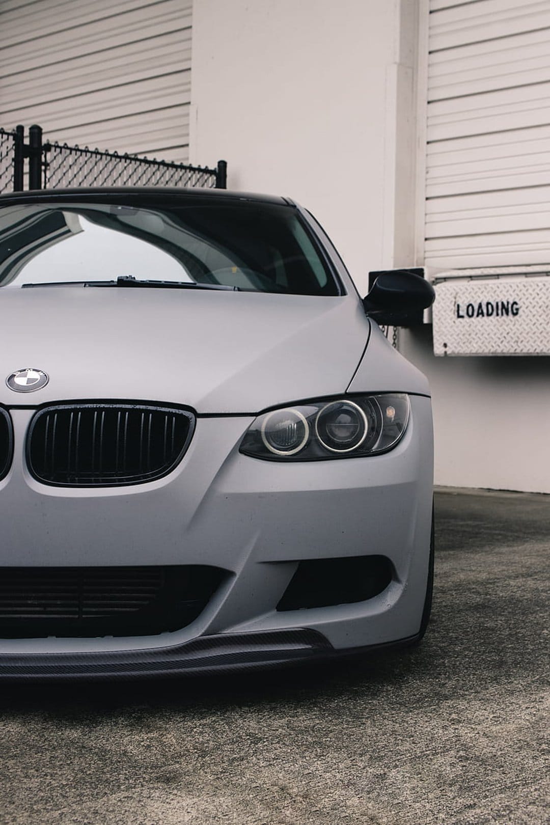 Matte gray BMW front end with a kidney grille and LED headlights parked by an industrial loading dock bay door.