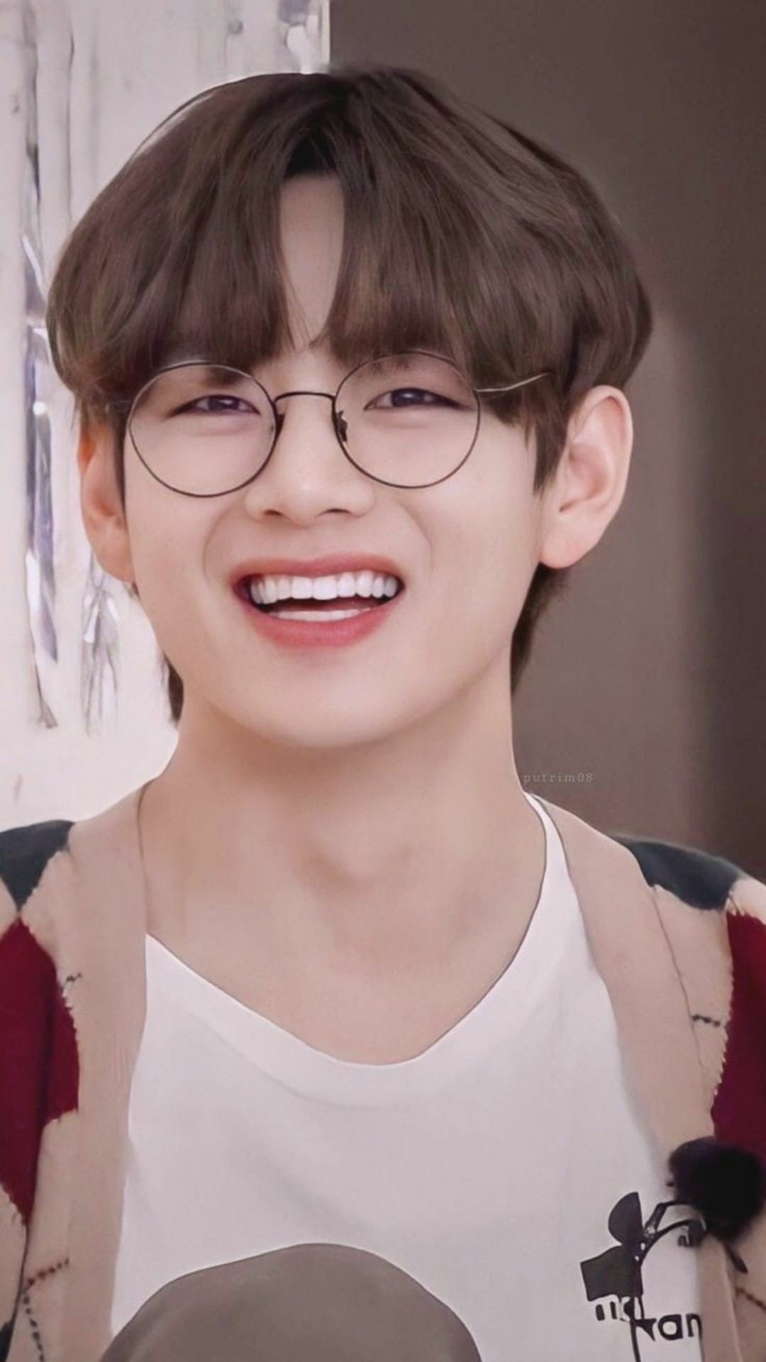 Kim Taehyung of BTS smiles broadly while wearing round glasses and a patterned cardigan in a close-up portrait.