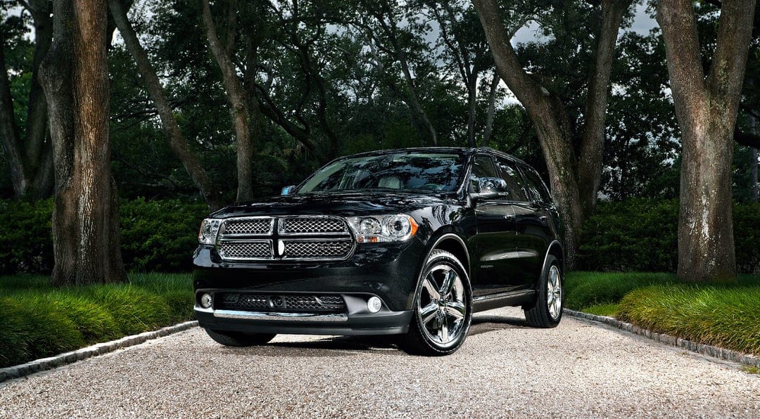 Black Dodge Durango SUV parked on a gravel path under tall green trees with bright daylight reflections.
