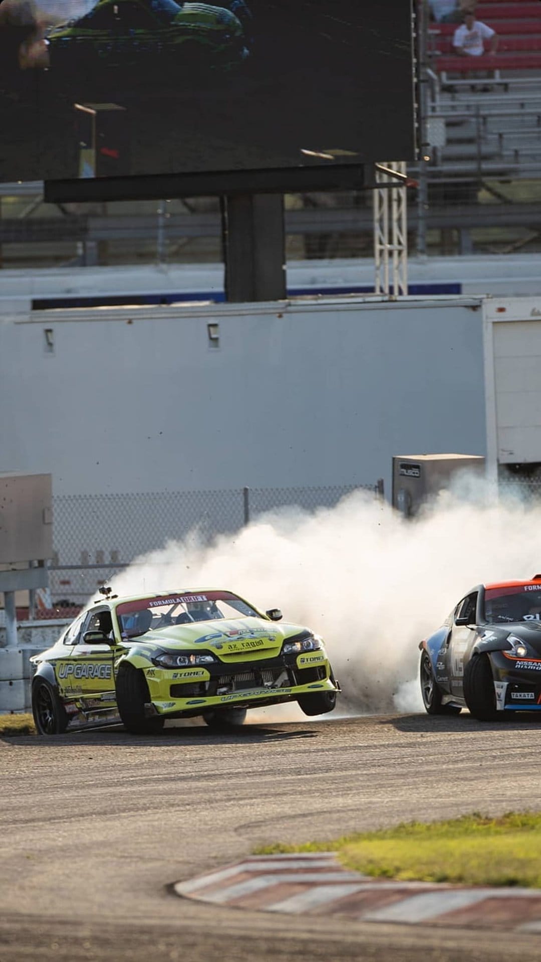 Race cars drift along a track with red and white curbs, producing a large white cloud of smoke in the sunlight.