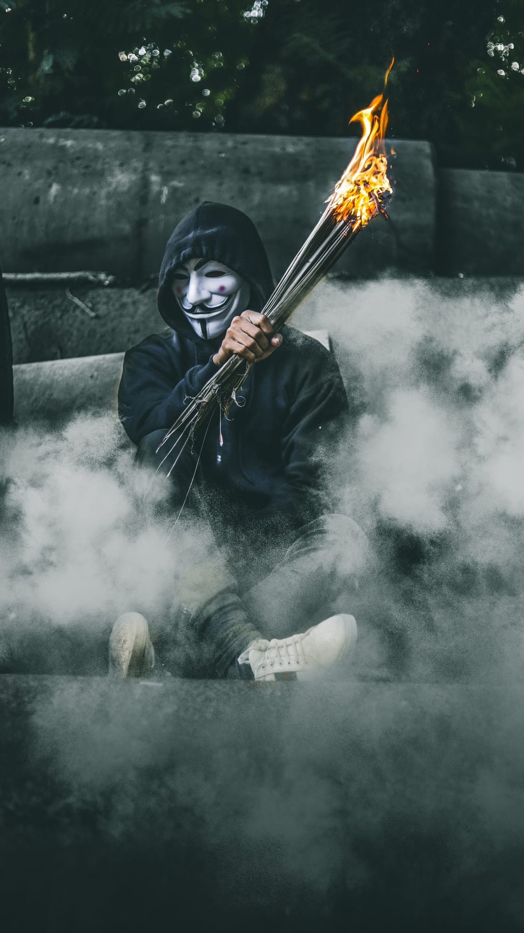 Masked person in a dark hoodie sits holding flaming sticks surrounded by dense white smoke in an urban setting.
