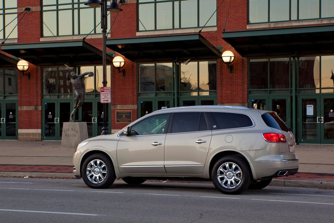 Champagne Buick Enclave SUV parked on an urban street in front of a red brick stadium with a bronze statue.