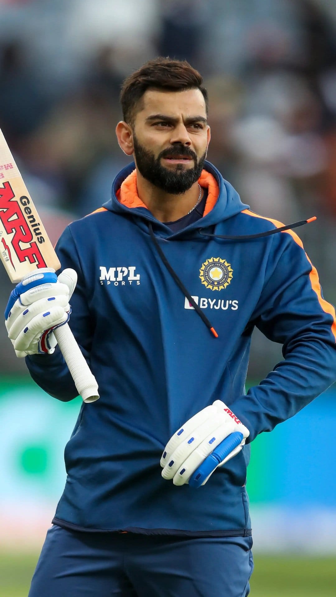 Virat Kohli wearing a blue Indian cricket hoodie and gloves while holding a bat against a blurred background.