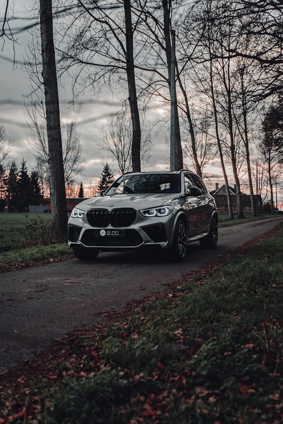 Silver BMW X5 M SUV on an asphalt road with bare trees and autumn leaves under a moody, overcast winter sky.