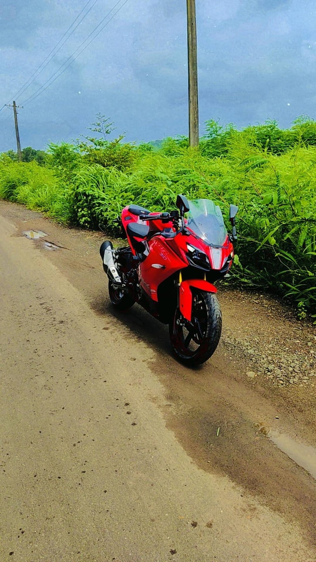 Red sport motorcycle parked on a wet unpaved road with dense green bushes under a dark stormy overcast sky.