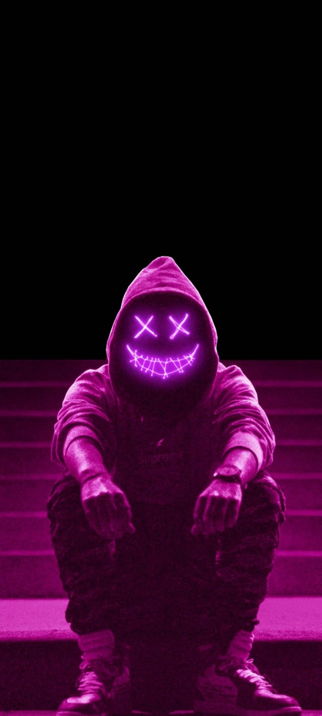 Person in a black hoodie wearing a glowing neon pink cross-eye mask while sitting on shadowy concrete stairs.