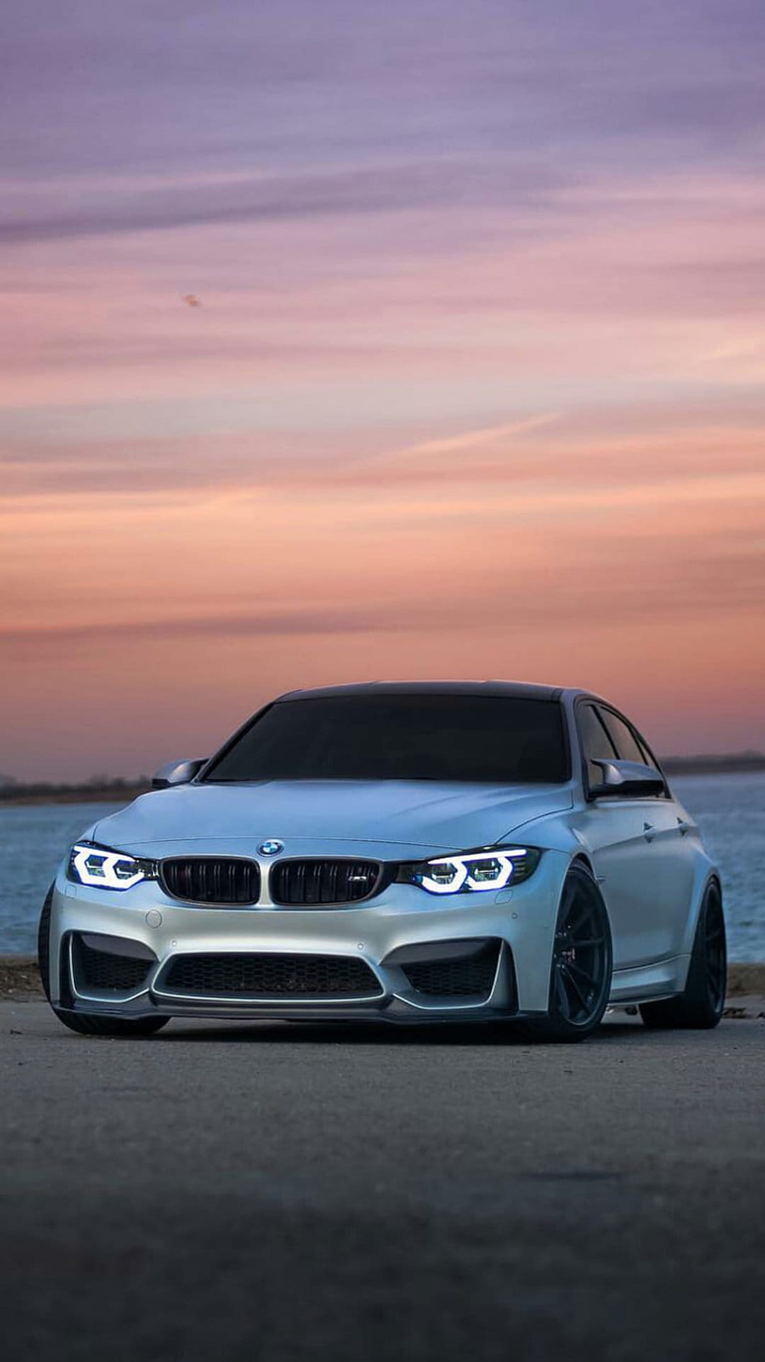 Silver BMW M3 coupe with glowing headlights parked by a purple sunset sky and calm waterfront at dusk.