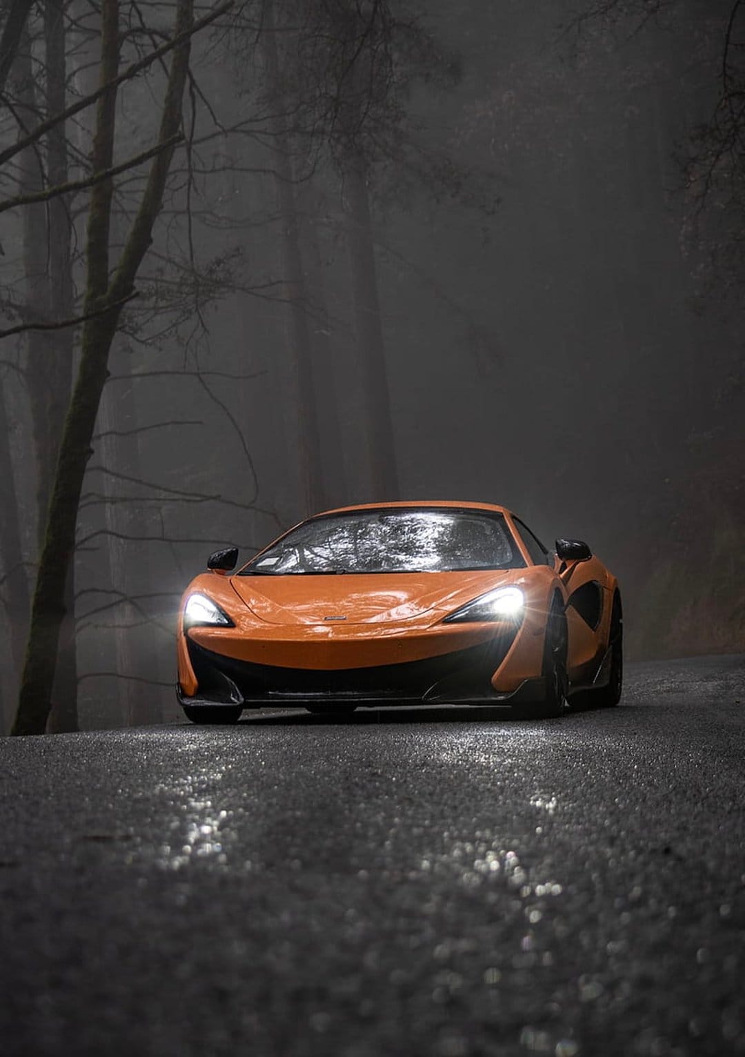 Orange McLaren 570S supercar driving on a wet road through a foggy forest with bare trees and glowing headlights.