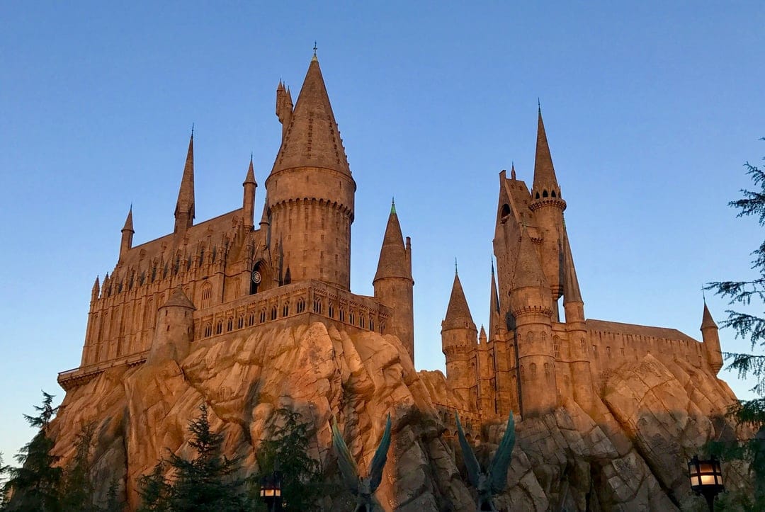 Hogwarts Castle towers glow with warm light against a dark blue evening sky atop a jagged rocky cliff.