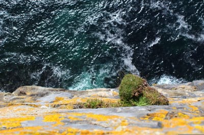 Weathered stone cliff with yellow lichen overlooks crashing blue waves and white sea foam in a vertical view.