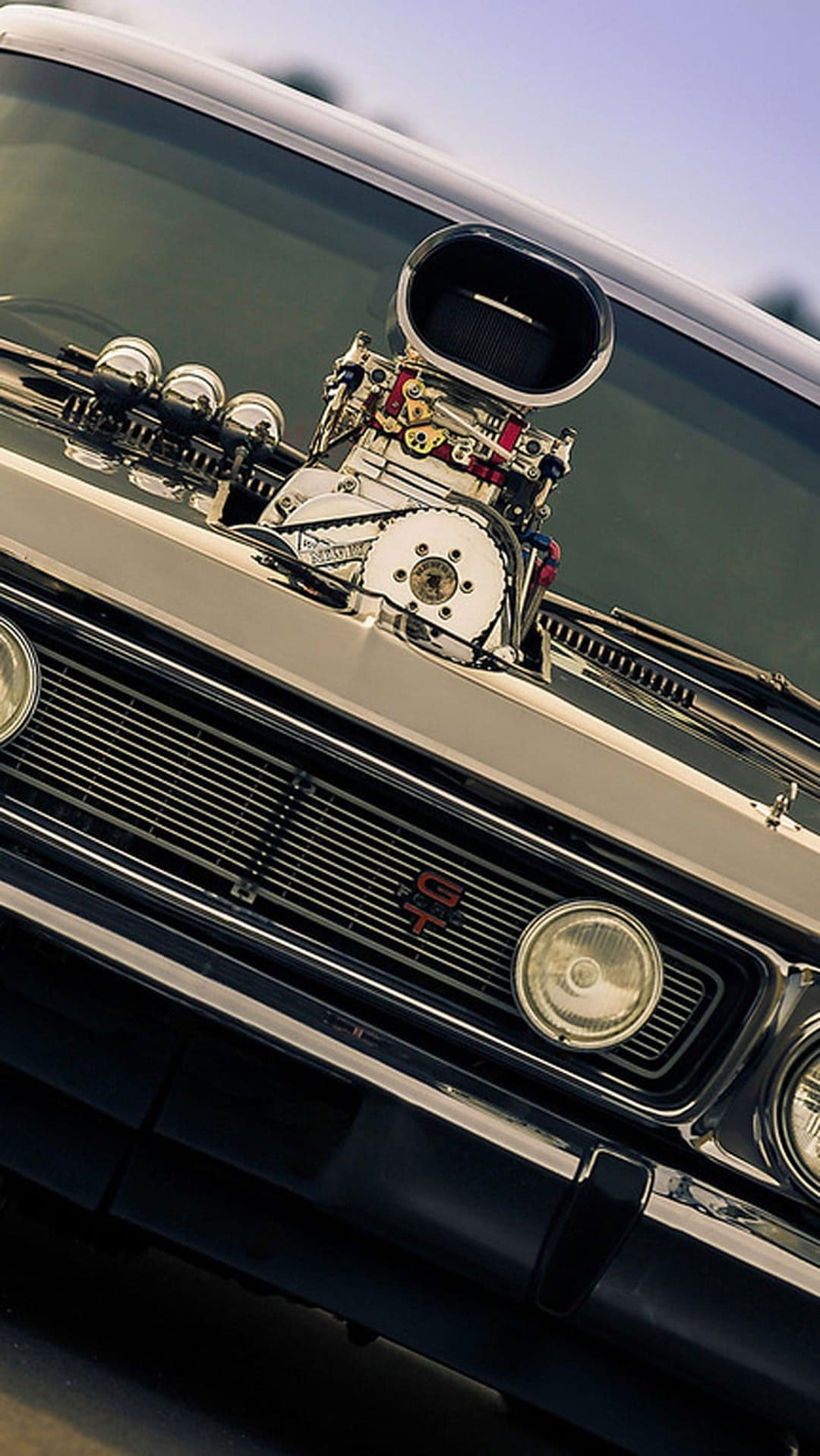 Chrome supercharger and carburetors rise from a vintage car hood beside a classic GT badge and front grille.