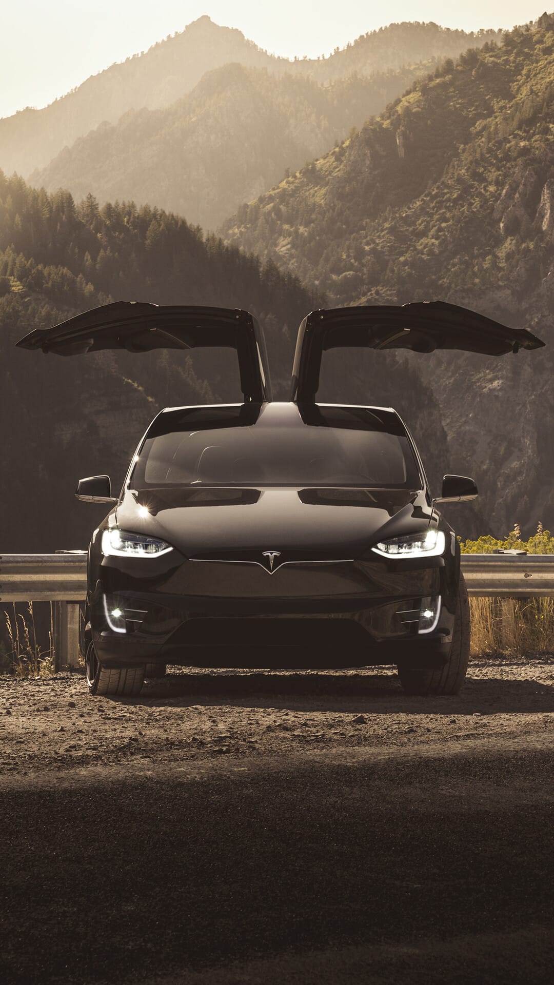 Black Tesla Model X with falcon wing doors open on a mountain road at sunset with glowing headlights.