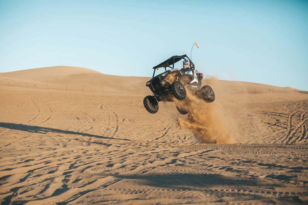 Side-by-side vehicle jumping over a sand dune with a massive dust cloud under a clear blue sky in bright sun.