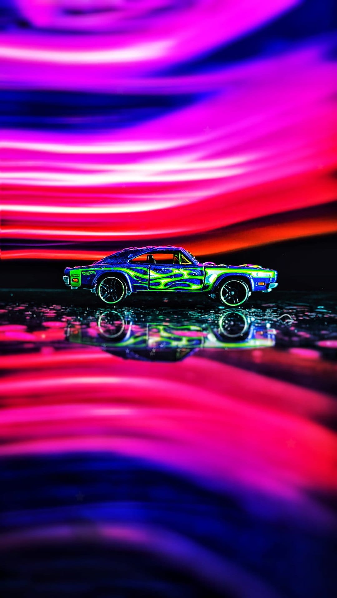 Blue toy car with green flames on a wet surface reflecting pink and purple neon light streaks in the dark.