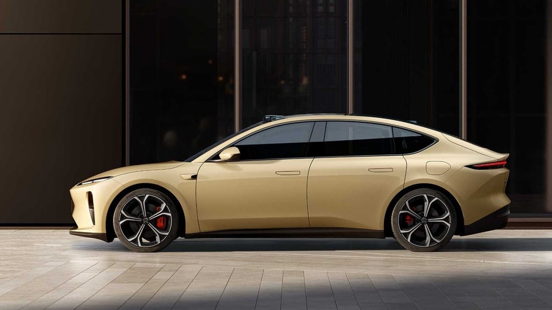 Champagne electric sedan side profile with black wheels and red calipers against a dark glass building.