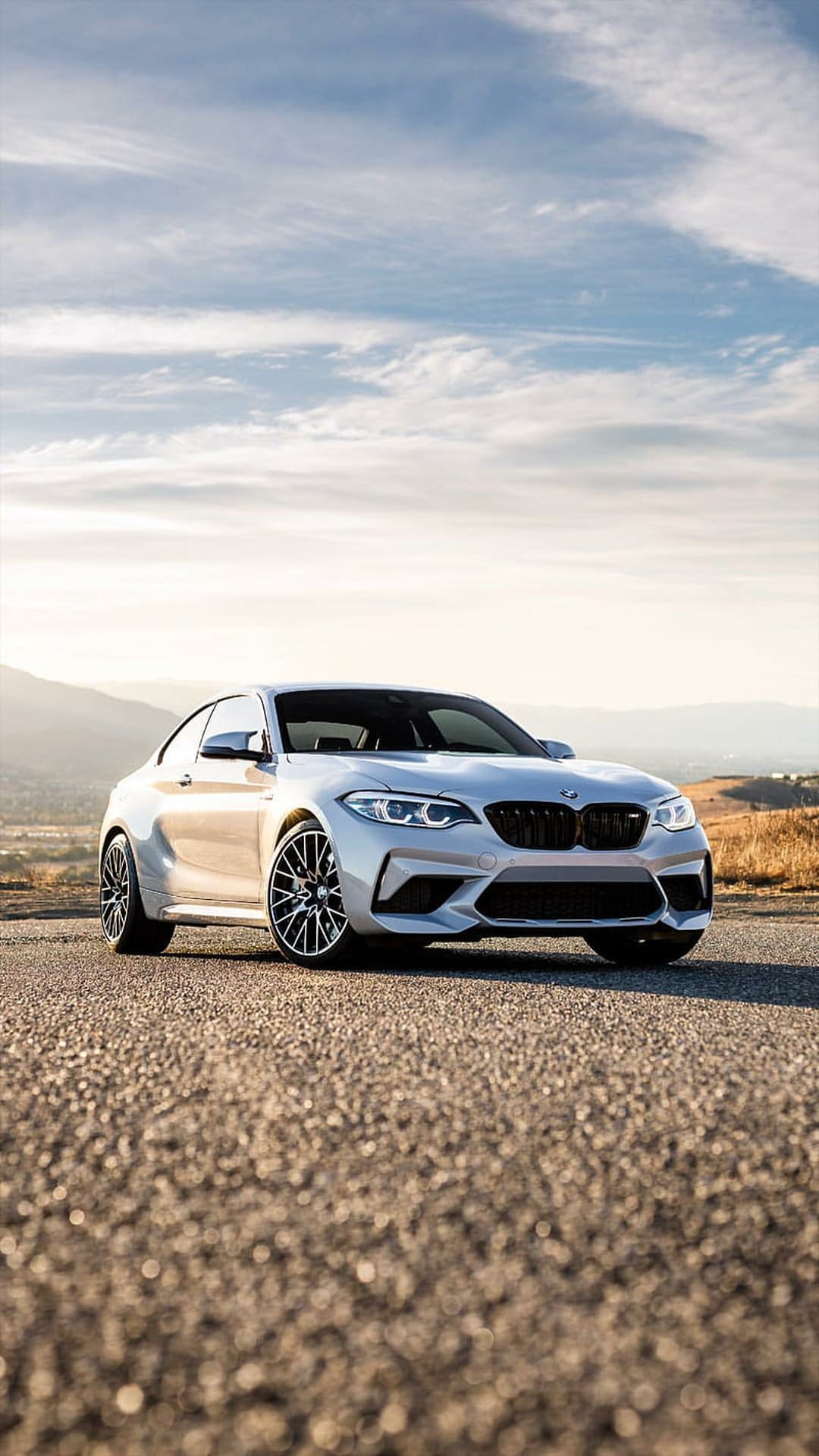 Silver BMW M2 Coupe parked on a gravel mountain road against a backdrop of distant peaks and a dramatic sky.