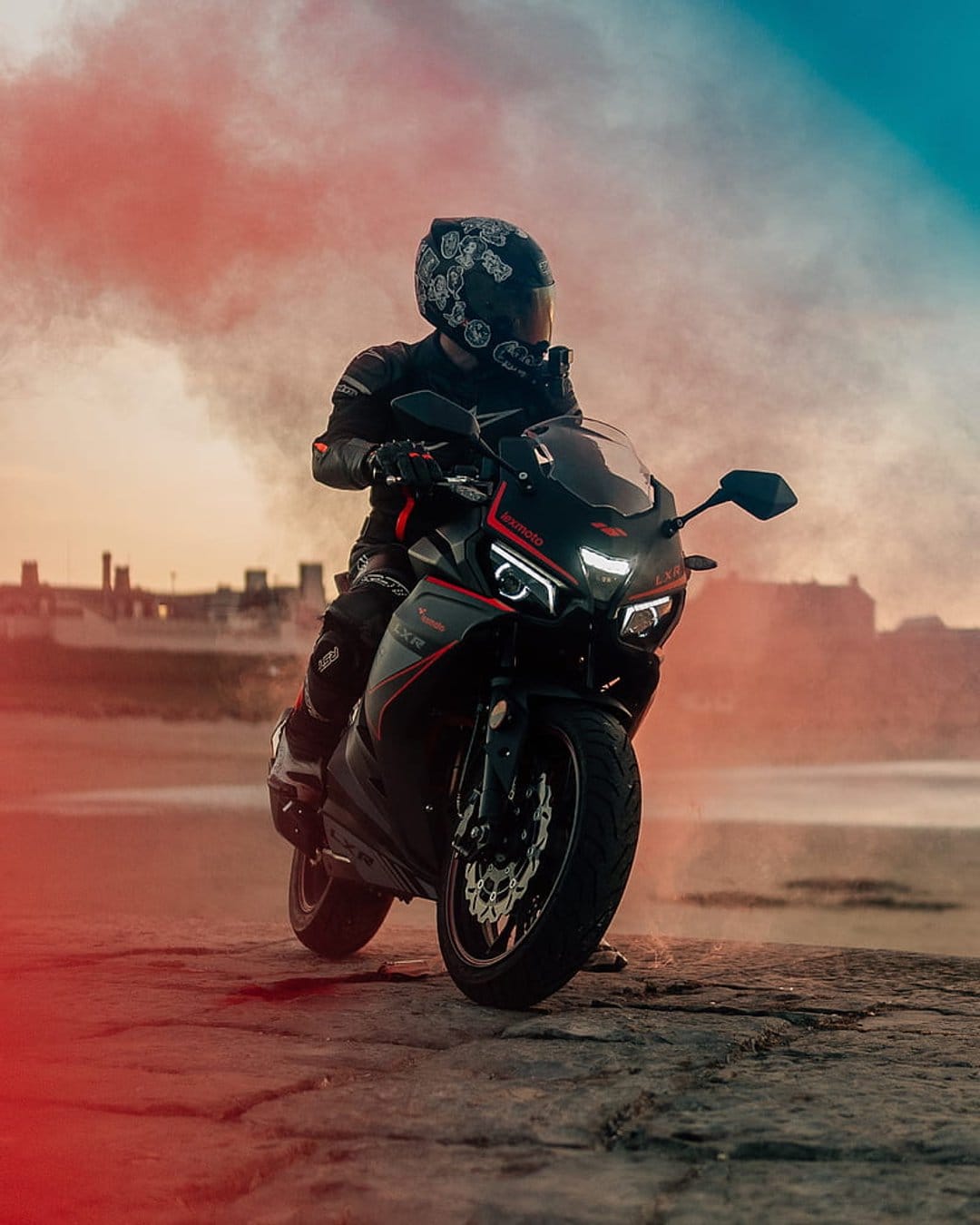 Biker in black armor sits on a sports bike amidst red smoke on a sandy beach with city buildings in the distance.
