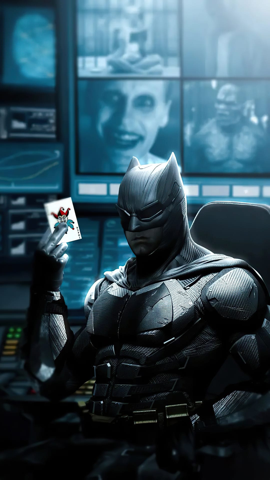 Batman in tactical armor holds a Joker card before glowing blue computer monitors in a dark high-tech cave.