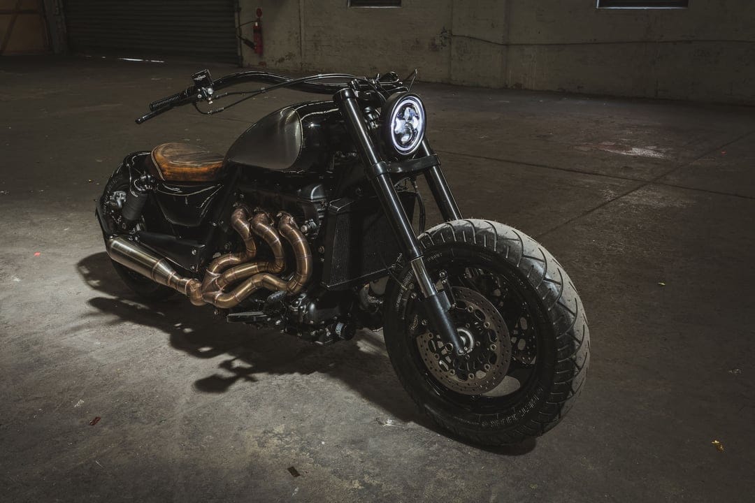 Black custom motorcycle with bronze exhaust pipes and a round LED headlight in a dark industrial warehouse.