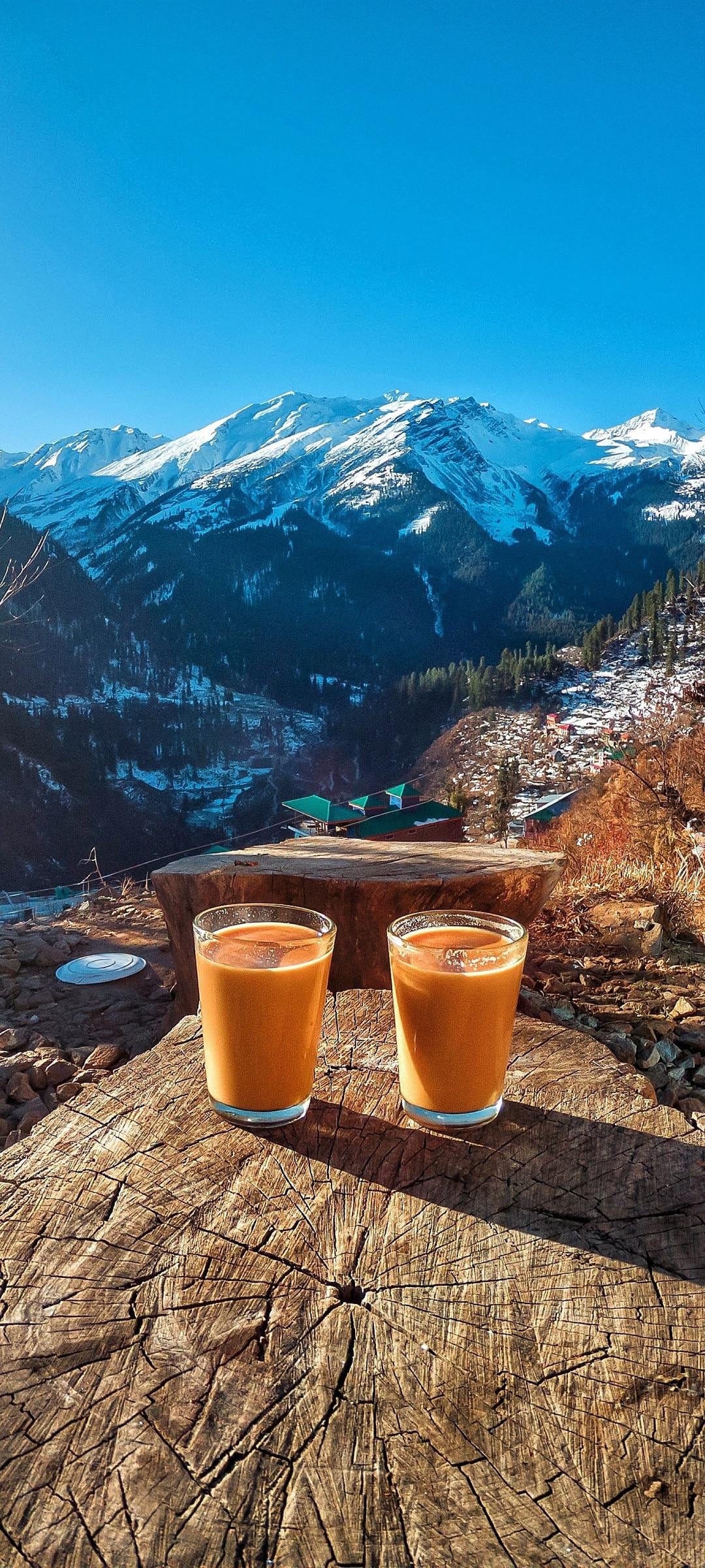 Two tea glasses on a wooden log overlook snowy mountain peaks and a green valley under a clear blue sky.