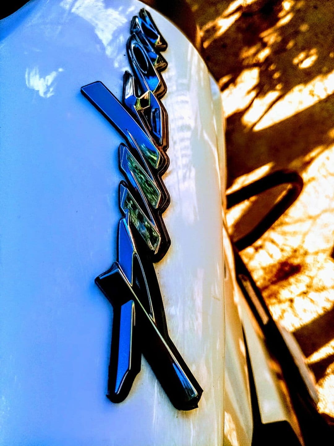 Chrome motorcycle emblem with blue accents reflecting sunlight and leaf shadows on a white metallic surface.