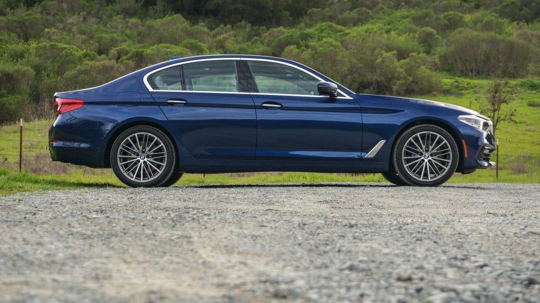 Blue BMW 5 Series sedan parked on a grey gravel path with green hills and trees in the background.