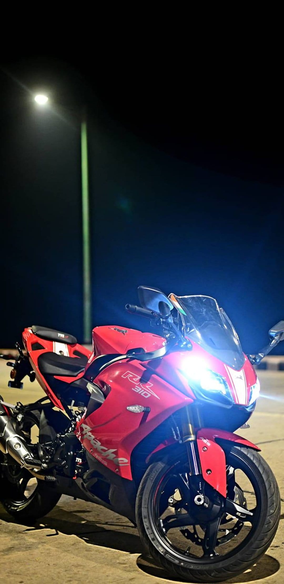 Red Apache RR 310 sportbike with glowing LED headlights parked on a dark road at night under streetlights.