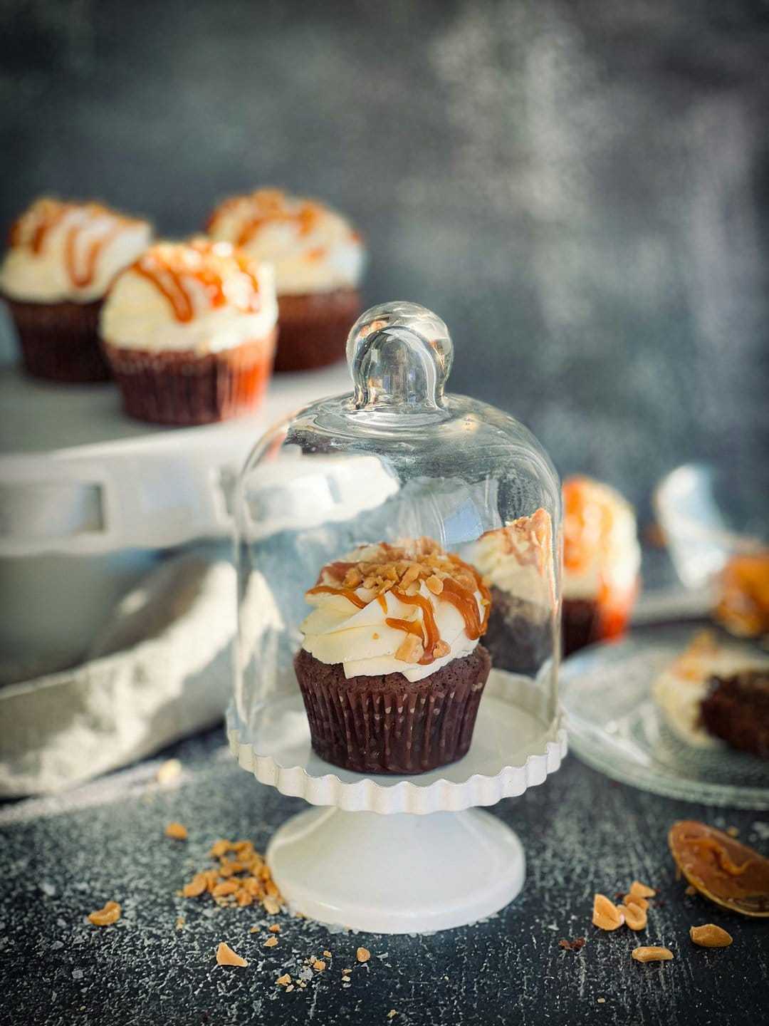 Chocolate cupcake topped with white frosting and caramel sauce sits under a glass dome on a white cake stand.