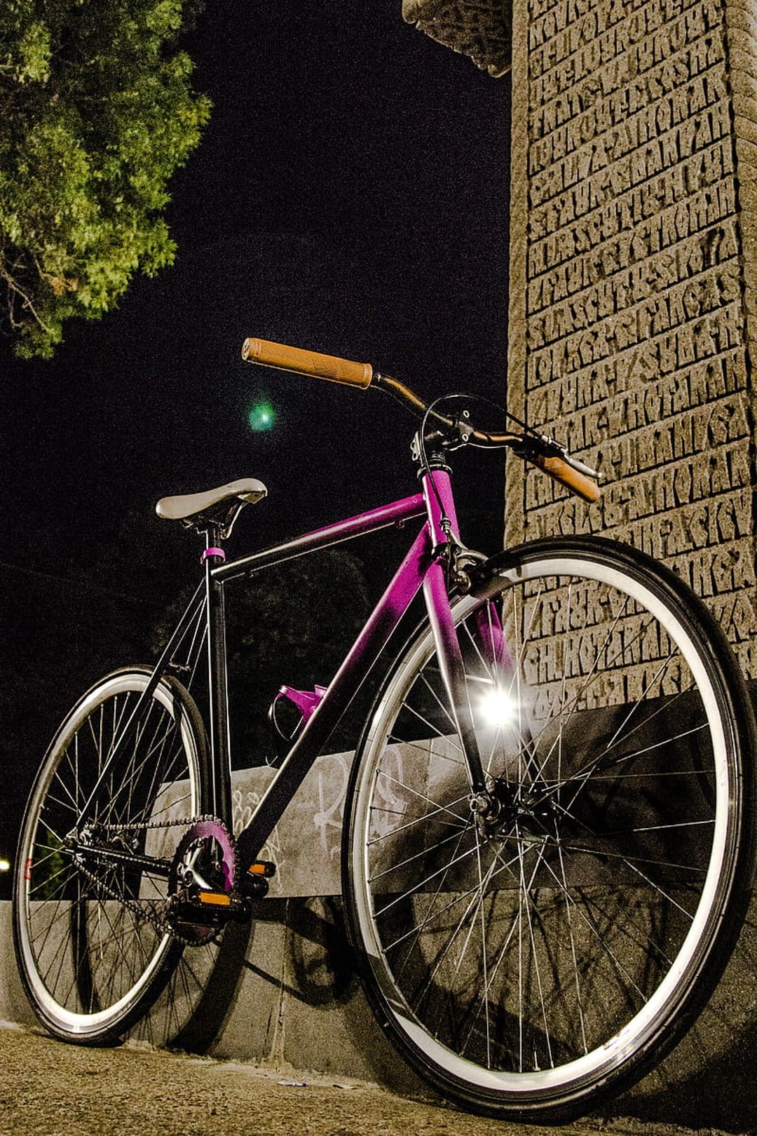 Purple and black fixed gear bike with a brown handlebar leaning against an inscribed stone wall at night.