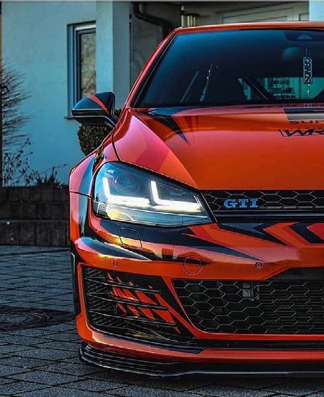 Orange Volkswagen Golf GTI with black racing stripes, glowing LED headlights, and a textured honeycomb front grille.
