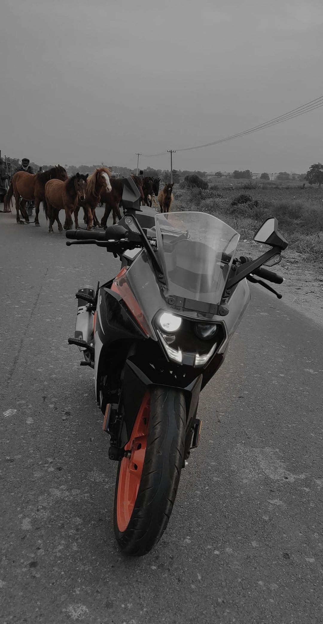 Orange sport bike on a rural road with riders on horseback in the background under a cloudy grey sky.