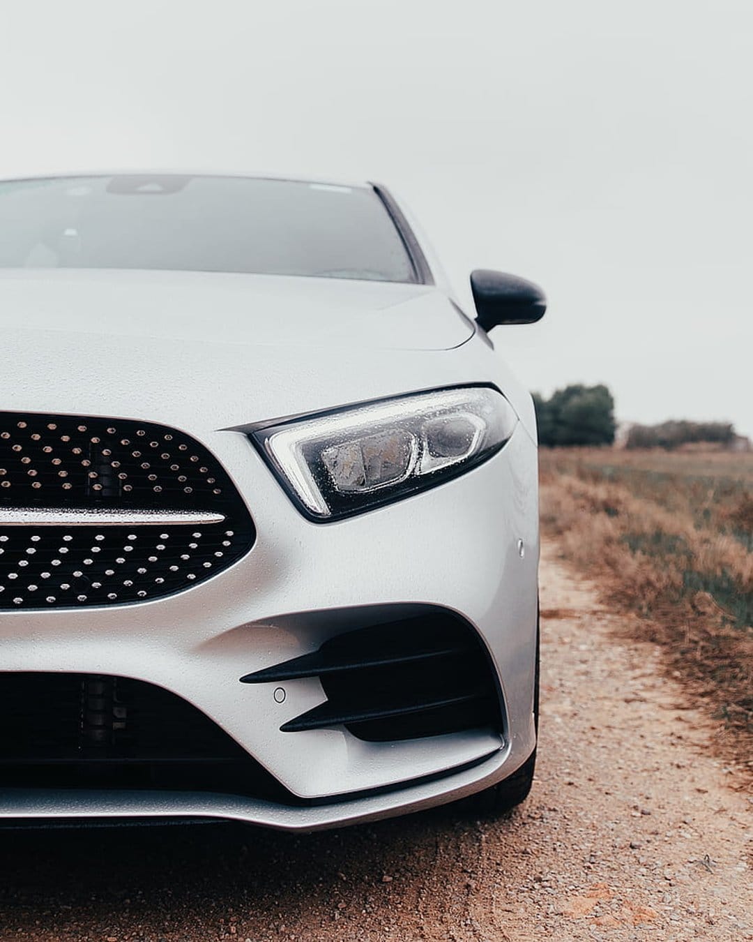 Silver car front grille and LED headlight on a gravel road with blurred trees under a gray overcast sky.
