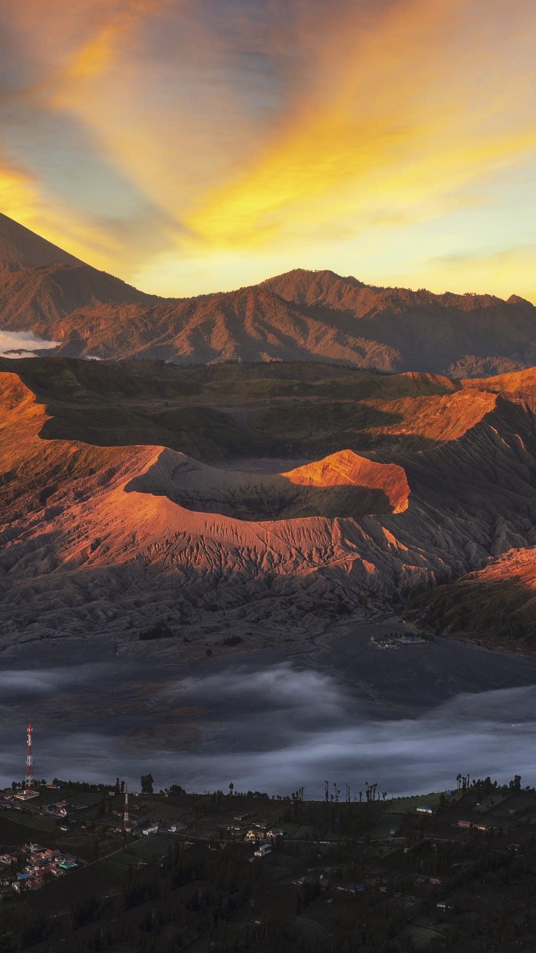 Mount Bromo peaks glow in golden light above a misty valley floor and a small Indonesian village at sunrise.