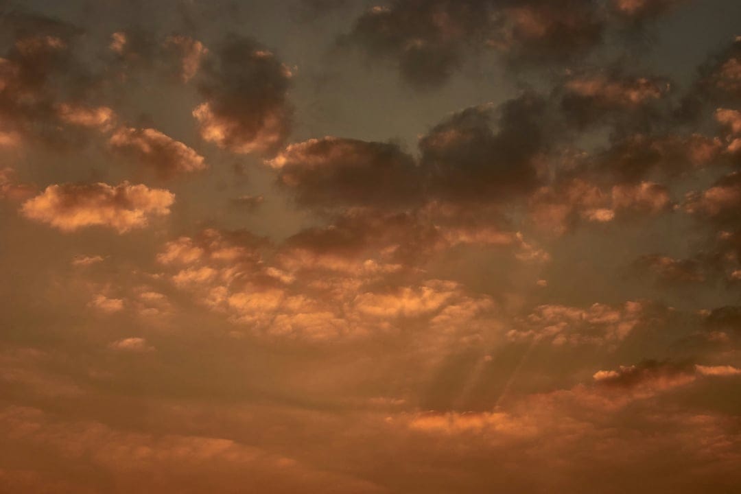 Orange clouds catch golden sunbeams in a hazy sky during a dramatic sunset with light rays piercing through.