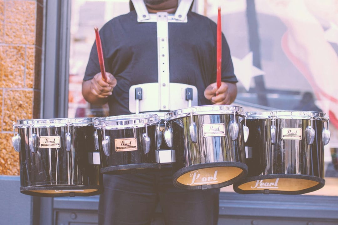 Marching band drummer in a black uniform holds red sticks above four black Pearl tenor drums outdoors.