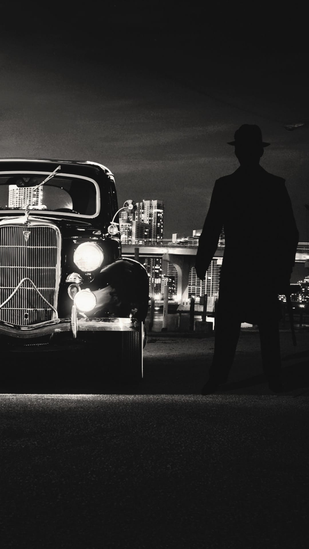 Silhouette of a man in a fedora standing by a vintage car with glowing headlights against a city skyline.