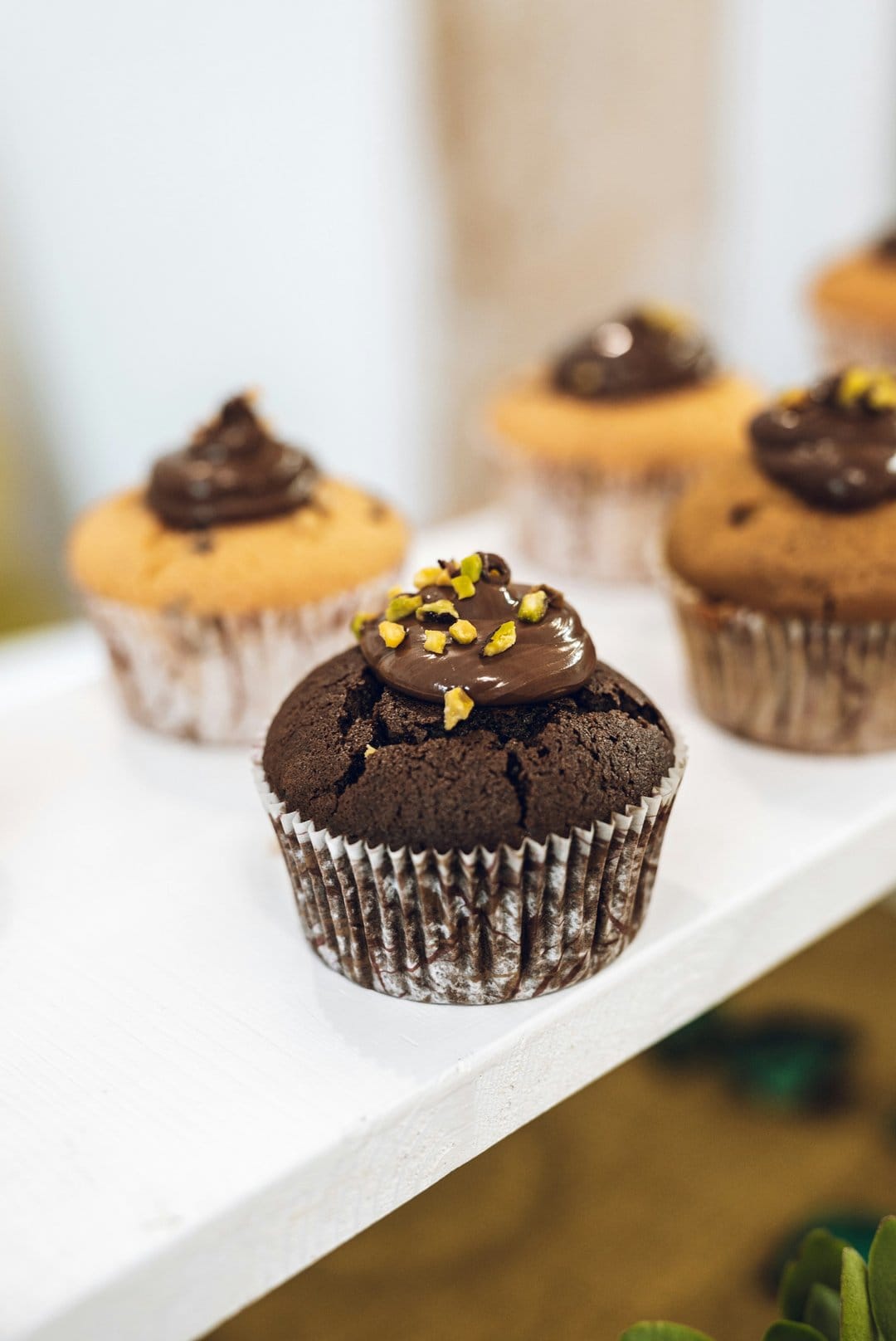 Chocolate cupcakes with swirls of dark frosting and crushed green pistachios against a blurry bakery backdrop.