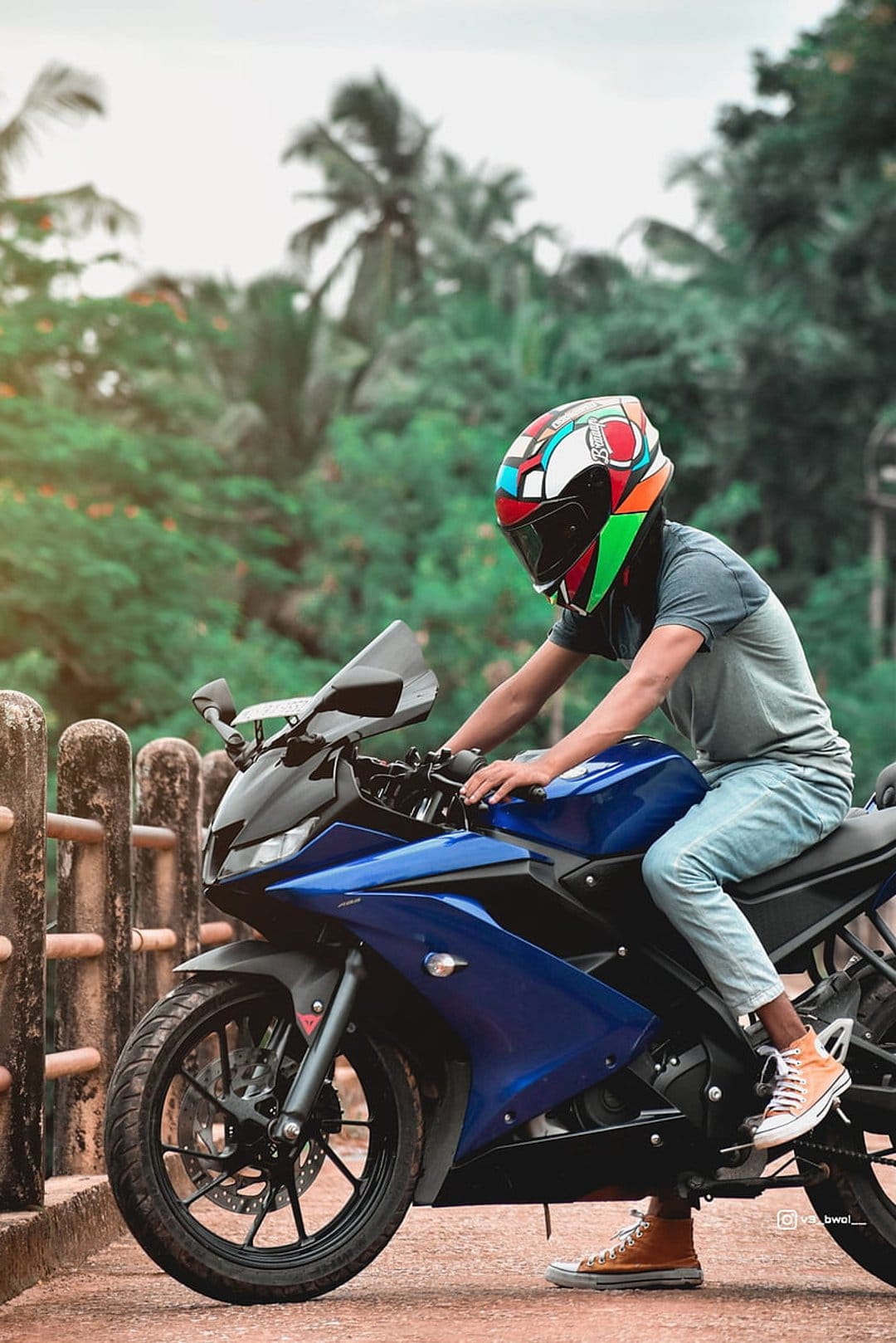 Young man on a blue motorcycle wearing a colorful helmet sits on a road surrounded by lush green forest trees.